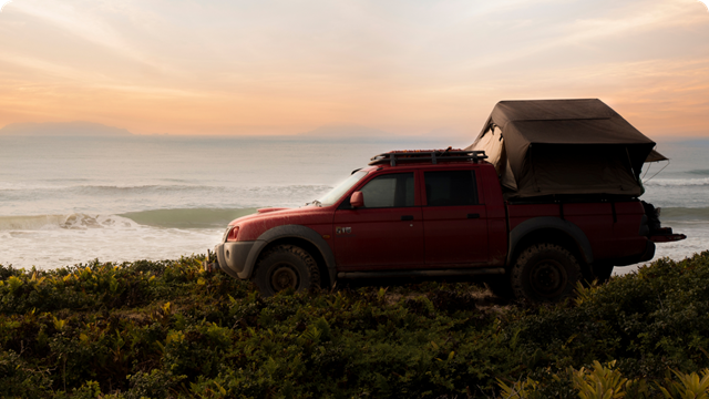 A red off-road vehicle with a rooftop tent parked on a grassy hill overlooking the ocean at sunrise.