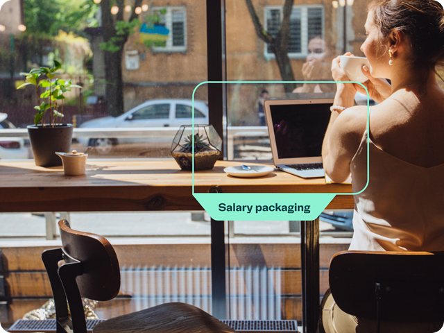 A woman sitting in a cafe near a large window, holding a cup of coffee. On the table in front of her is a laptop and a small plant. The image includes a green label with the text 'Salary packaging' highlighting the laptop. 