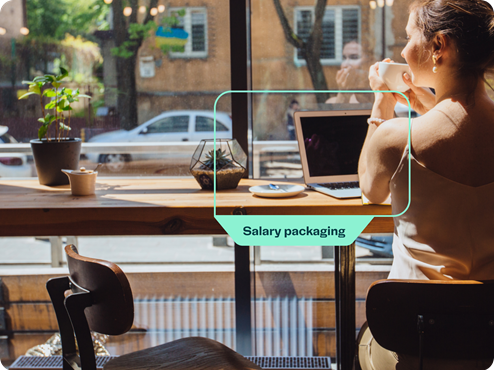 A woman sitting in a cafe near a large window, holding a cup of coffee. On the table in front of her is a laptop and a small plant. The image includes a green label with the text 'Salary packaging' highlighting the laptop. 