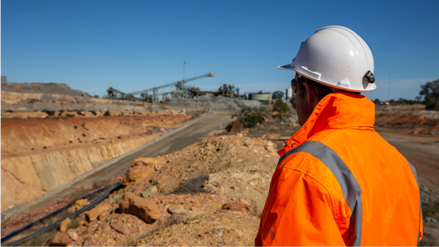 A miner dressed in an orange high-visibility jacket and hard hat surveys an open pit, highlighting the mining operation.