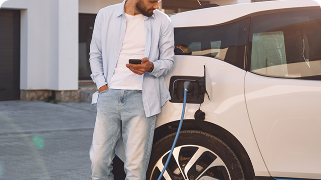 A car owner standing while holding their mobile phone in front of a white electric vehicle as it charges. 