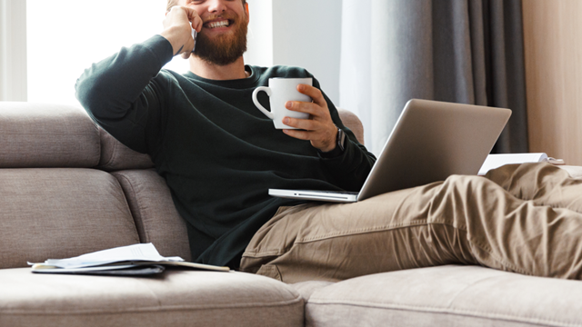 A person reclining on a lounge using multiple portable elctronic devices, talking on a mobile phone while a laptop rests on their lap. 