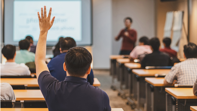 A classroom setting with a student raising their hand while a person at the front is teaching. 