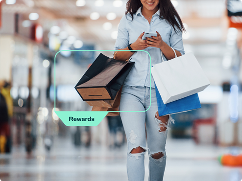 A person walking in a mall holding shopping bags and a smartphone with a 'Rewards' label highlighting their shopping.